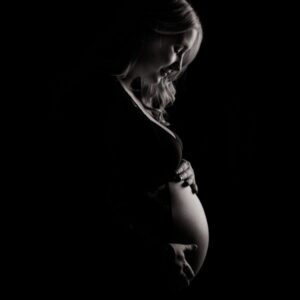 A woman standing in the dark holding her pregnant belly.